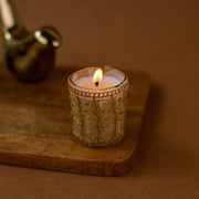Lit decorative candle in gold glass holder on wooden tray, brown background
